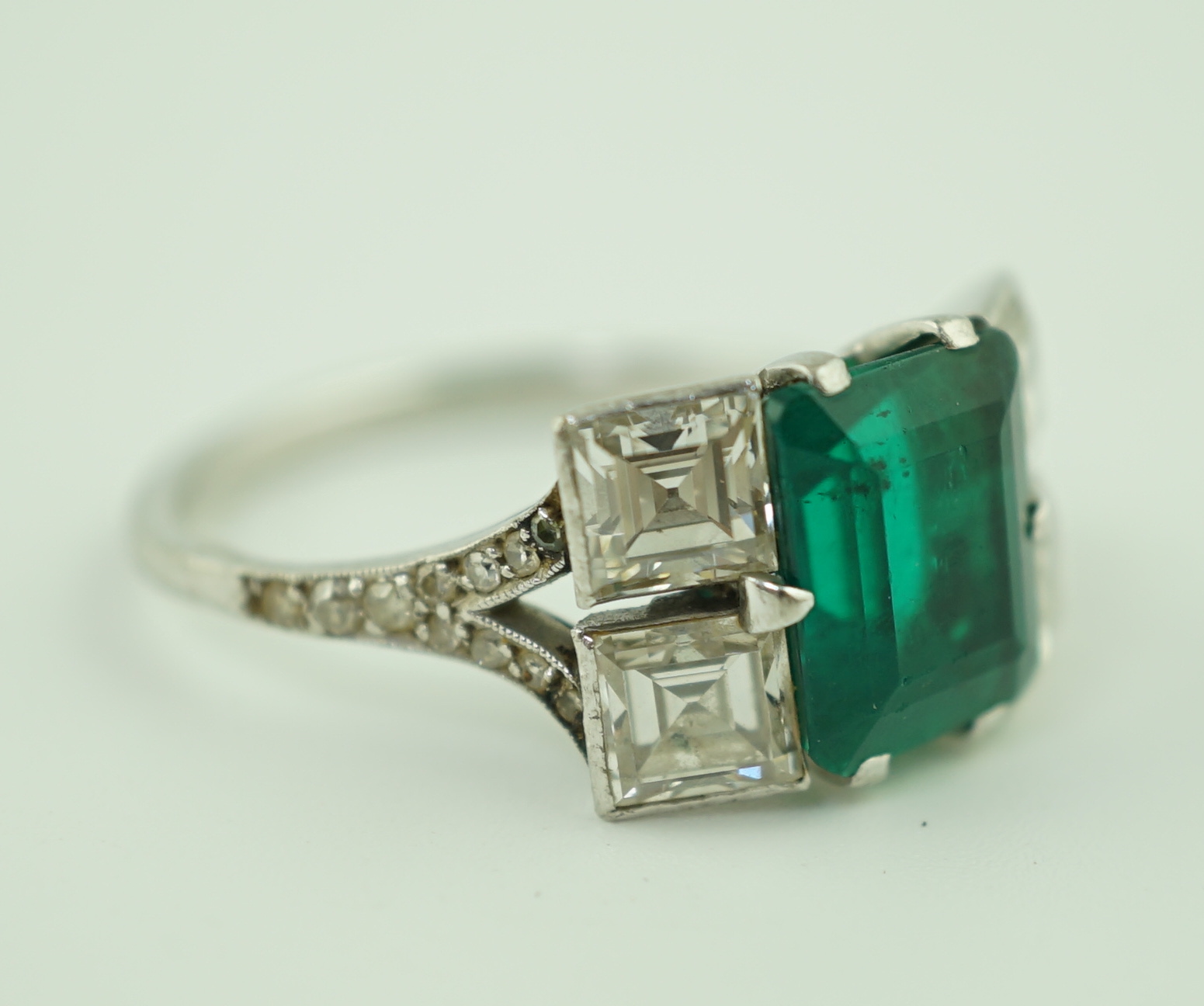 An Art Deco platinum, emerald and square cut diamond dress ring, with diamond chip set shoulders